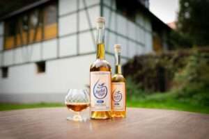 Bild mit einer großen und einer kleinen Flasche Apfeltau Fusion sowie einem halbgefüllten Glas, die auf einem Tisch im freien stehen